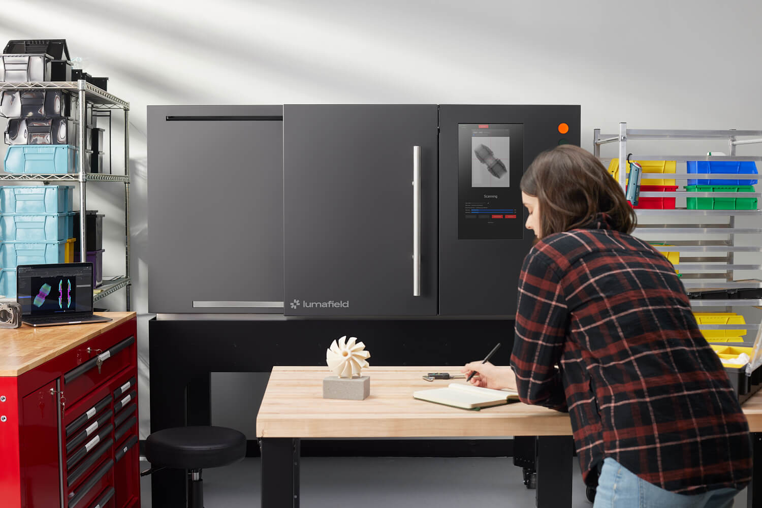 A person stands at a wooden workbench with a notepad, looking at a 3D scanner in a well-organized workspace. The machine displays an image of a scanned object. The background includes shelves with colorful bins, storage boxes, and tools.