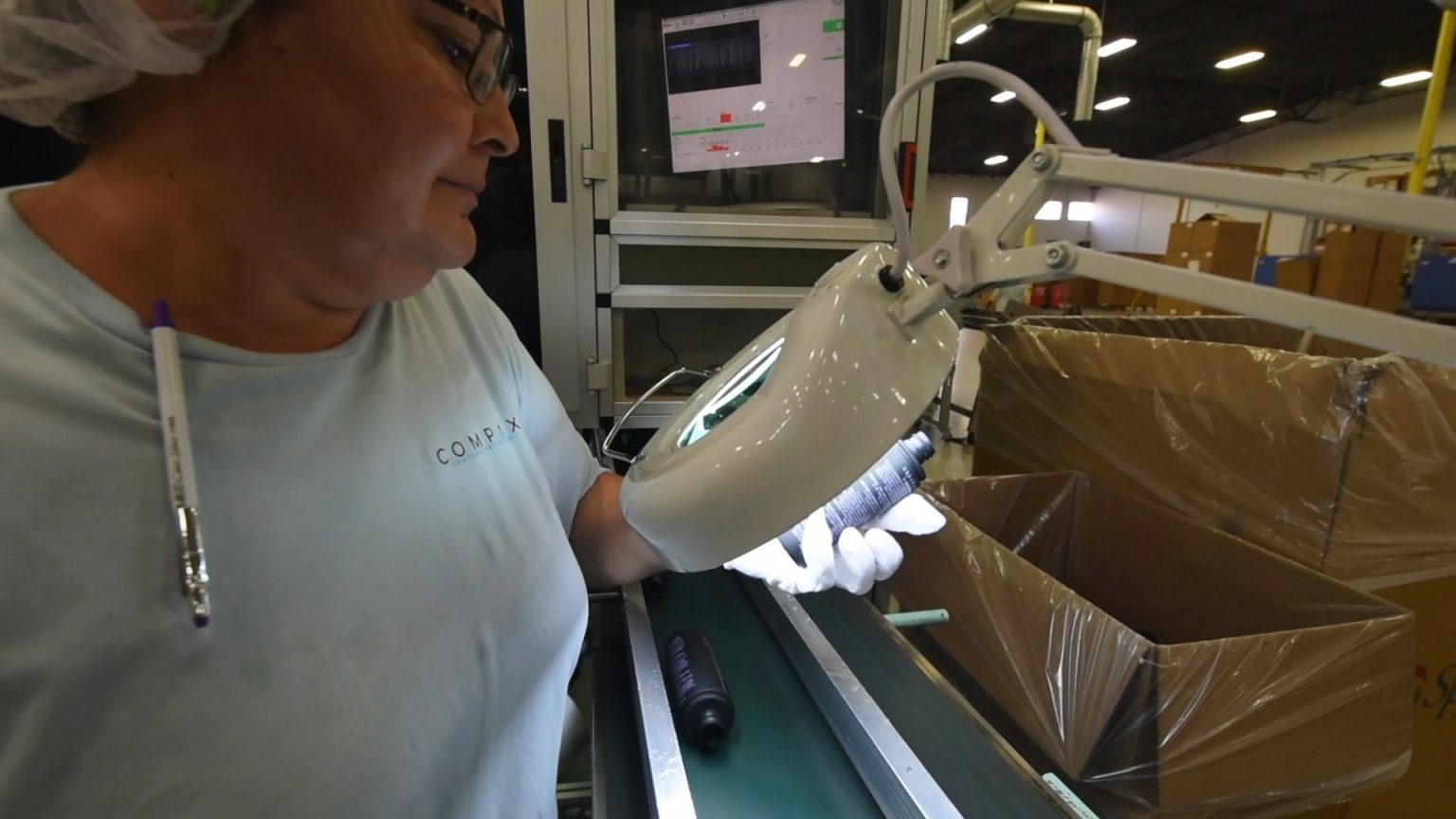 A person wearing glasses and a hairnet inspects a small handheld item under a lit magnifying lamp on a conveyor belt in a factory. A computer screen and large cardboard boxes are visible in the background.