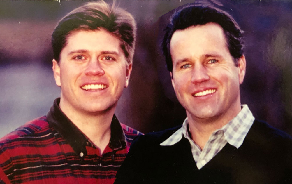 Two men are smiling and standing close together. The man on the left is wearing a red plaid shirt, while the man on the right is wearing a dark sweater over a checkered shirt. The background is blurred, highlighting the men as the main focus of the image.