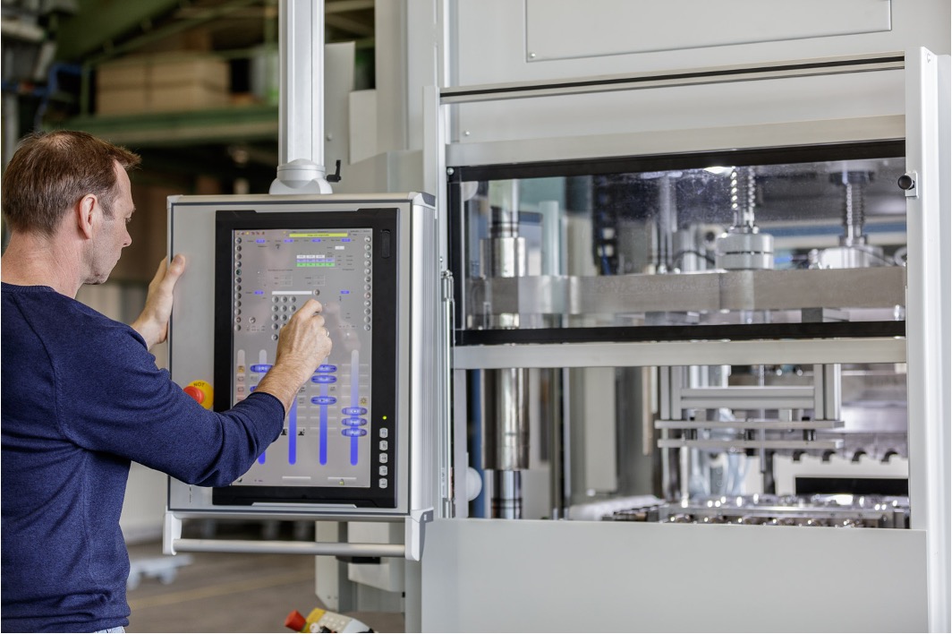 A person operates a large piece of industrial machinery using a touchscreen interface. The machinery has multiple components, including metal parts and a large compartment with various elements visible through a glass panel.