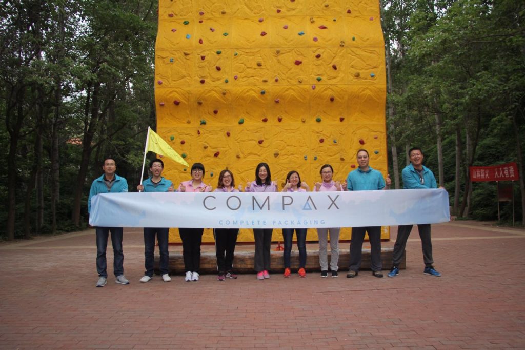 A group of nine people is standing outdoors in front of a large yellow climbing wall. They are holding a long banner that reads "COMPAX COMPLETE PACKAGING." The group is smiling and dressed in casual attire, with some wearing jackets and others in t-shirts.