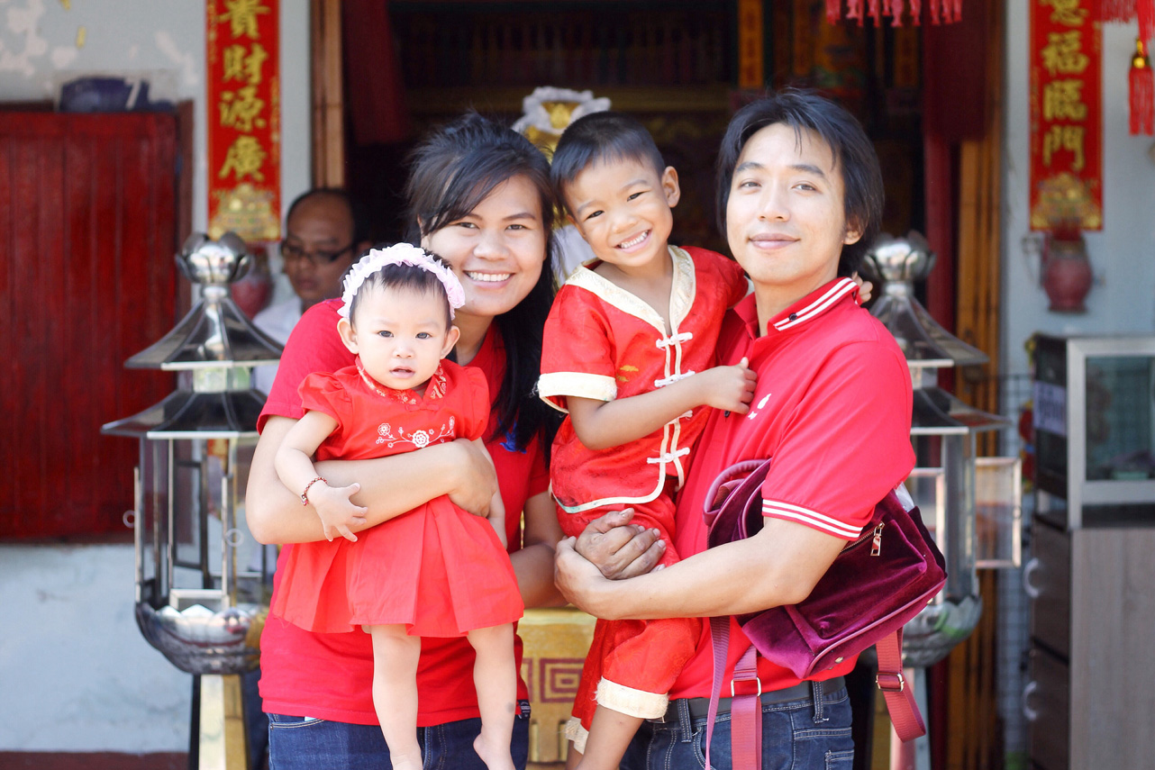 A family of four, dressed in red, stands smiling in front of a traditional Chinese building. The mother holds a small girl in a red dress, while the father carries a young boy in a matching red outfit. The parents wear red tops, embracing their children warmly.