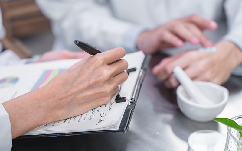 Close-up of two individuals working together around a table. One person is writing on a clipboard with a pen, and the other is holding a mortar and pestle. The setting appears to be a laboratory or a clinical environment.