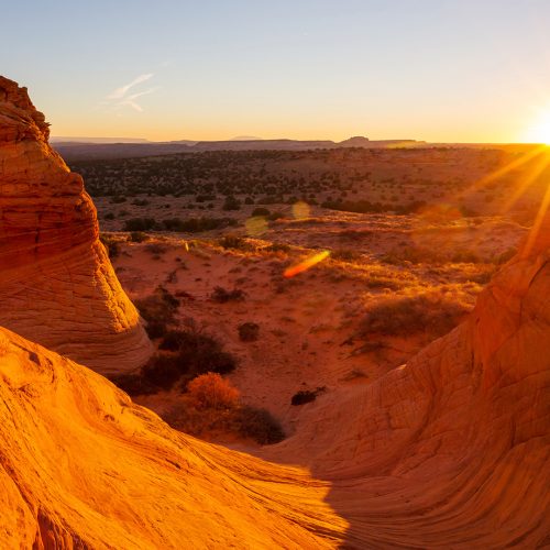 A stunning desert landscape at sunset with rocky formations illuminated by the warm, golden glow of the setting sun. The sun is partially visible on the horizon, casting shadows and light across the sandy terrain and distant flatlands.